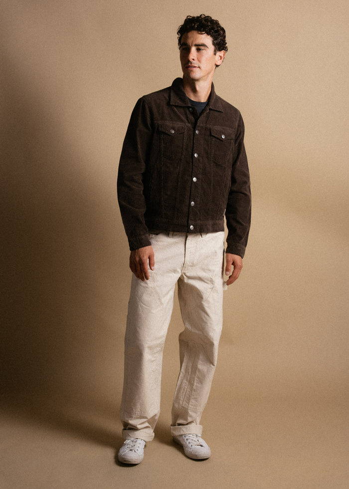 Man in brown jacket and beige pants stands against a tan background, looking to the side with a relaxed posture and expression.