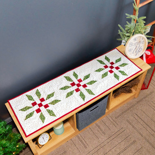 A Christmas-themed quilted bench runner with three green and red snowflake patterns on a white background, styled on a wooden bench in a decorated room.