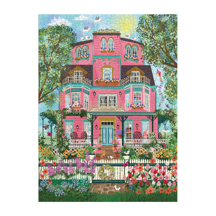 The Joy Laforme Capeside Victorian 1000 Piece Foil Puzzle features a vibrant pink Victorian mansion with balconies, blooming flowers, lush gardens, and a blue sky backdrop—artwork by Joy Laforme.
