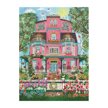 The Joy Laforme Capeside Victorian 1000 Piece Foil Puzzle features a vibrant pink Victorian mansion with balconies, blooming flowers, lush gardens, and a blue sky backdrop—artwork by Joy Laforme.