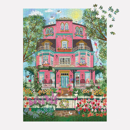 The Joy Laforme Capeside Victorian 1000 Piece Foil Puzzle depicts a grand Victorian mansion with balconies, trees, a white picket fence, and flowers. A few pieces remain unfinished in the upper right corner. Brand: Joy Laforme.